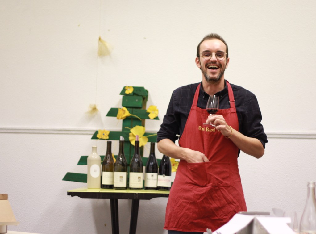 Jeremy Debreu souriant, un verre de vin à la main, devant un sapin de Noël en papier et une rangée de bouteilles