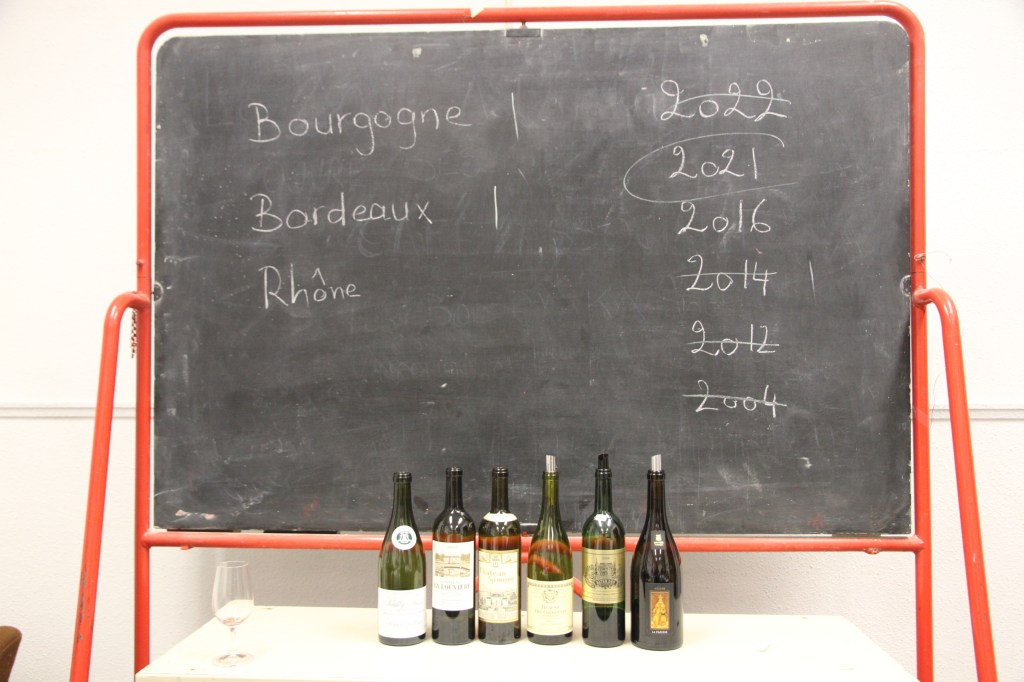 Tableau noir listant Bourgogne, Bordeaux et Rhône avec des millésimes, bouteilles posées au pied