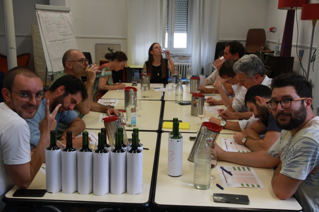 Équipes autour de tables de dégustation à l’aveugle, bouteilles habillées de papier et fiches de notation